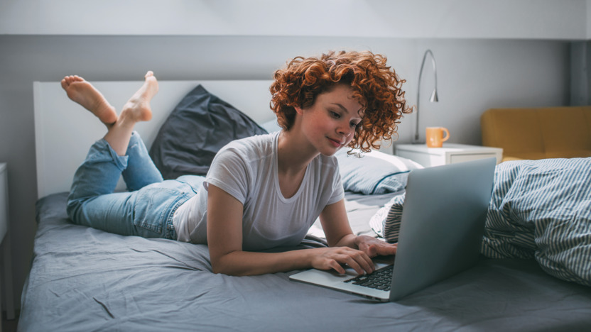 Junge Frau mit rotem Lockenkopf liegt auf dem Bett und surft mit einem Laptop im Internet