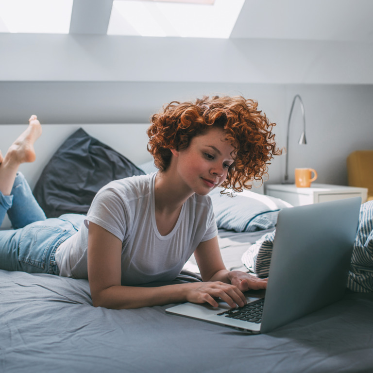 Rothaarige Frau liegt im Bett vor dem Laptop. 