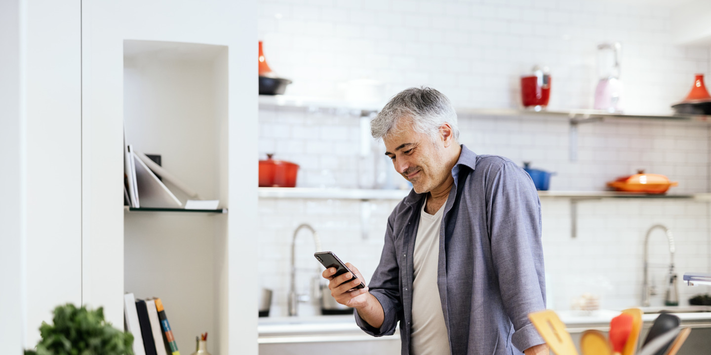 Grauhaariger Mann steht in Küche und hält Smartphone in der Hand. 