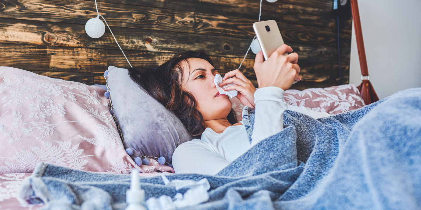 Frau liegt krank mit Schnupfen im Bett und betätigt ihr Smart Phone