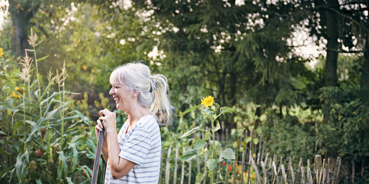 Grauhaarige Frau steht in Garten und hält Schaufel in der Hand. 