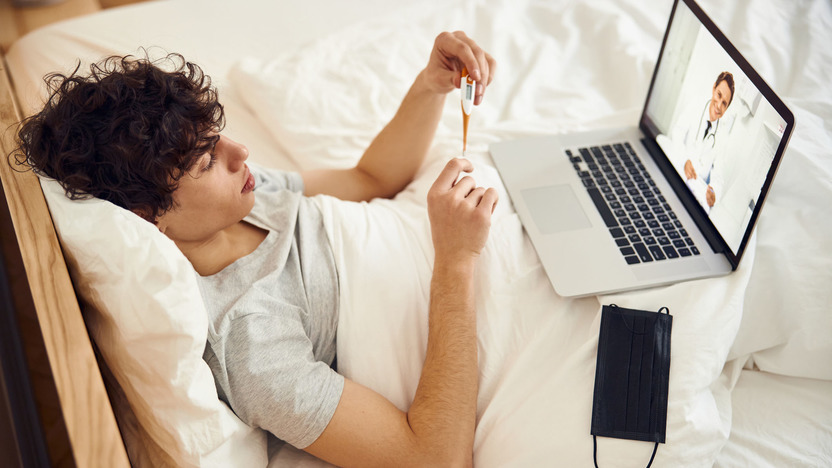 Junger Mann liegt mit einem Fieber-Thermometer und Laptop im Bett
