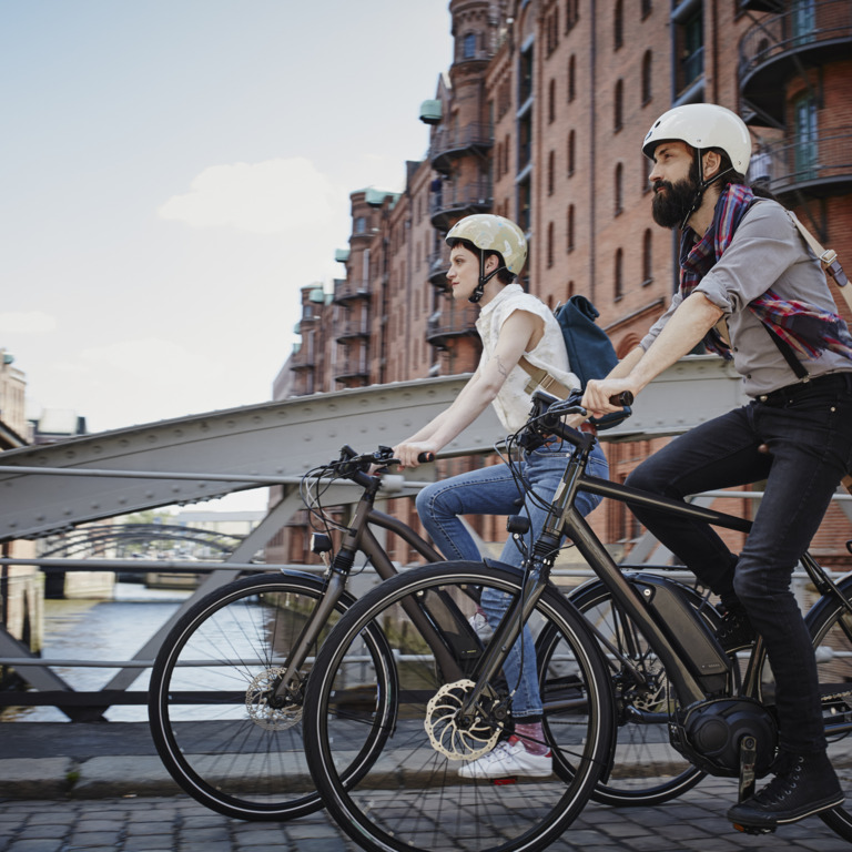 Junge Man und Frau fahren mit dem Fahrrad über die Brücke. Sie sind leicht angezogen und beide tragen einen Helm.