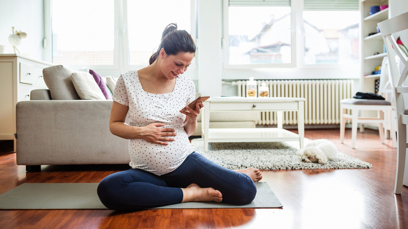 Schwangere Frau sitzt auf Yoga-Matte und tippt auf ihrem Smartphone