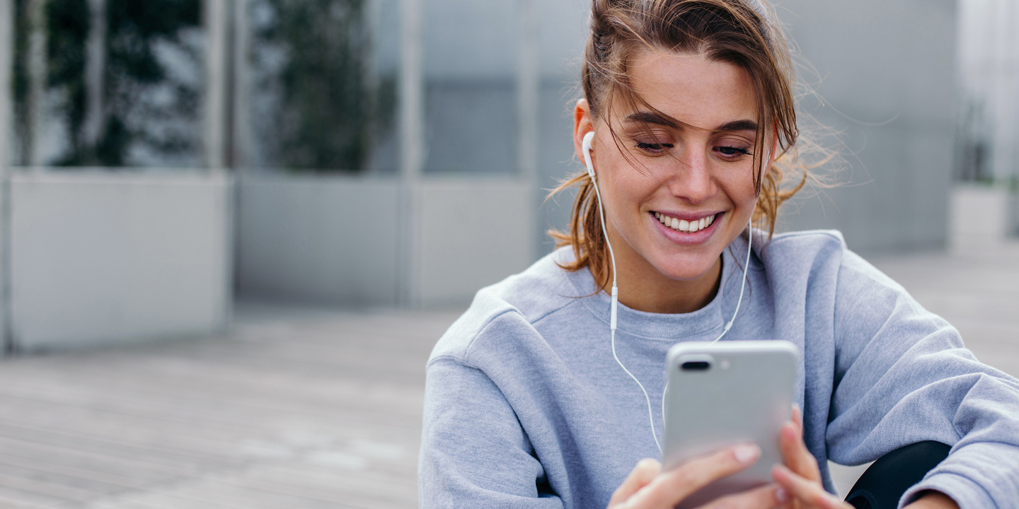Junge Frau sitzt draußen und hört über Kopfhörer auf ihrem Handy einen Podcast