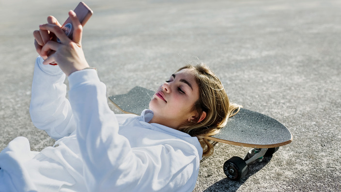 Junges Mädchen stützt ihren Kopf auf einem Skateboard und hält dabei ihr Handy in der Hand. 