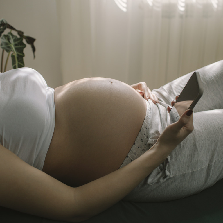 Schwangere Frau liegt auf Bett und schaut auf ihr Smartphone.