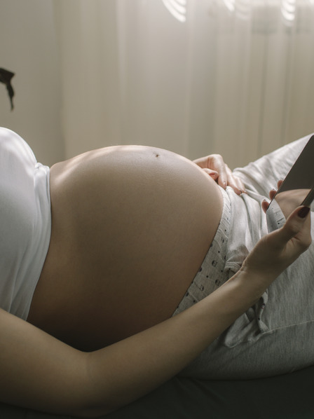 Frau liegt schwanger auf dem Bett und schaut in ihr Handy.