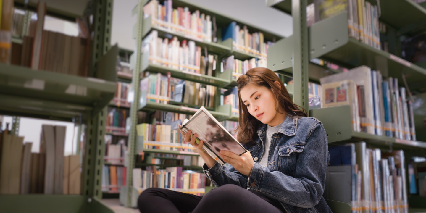 Junge Frau sitzt mit aufgeschlagenen Buch in der Hand auf dem Boden vor vollen Bücherregalen