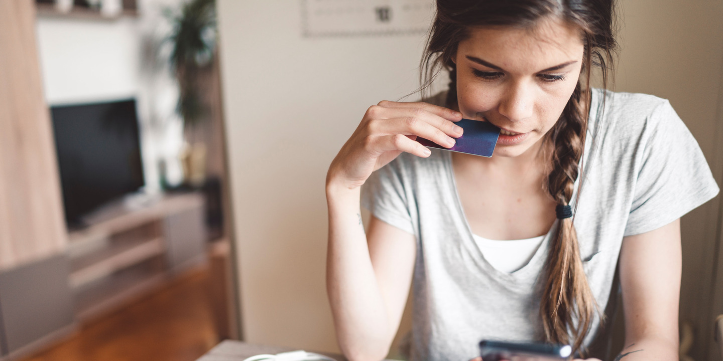 Junge Frau beißt leicht in ihre Kreditkarte und schaut unglücklich auf ihr Handy.