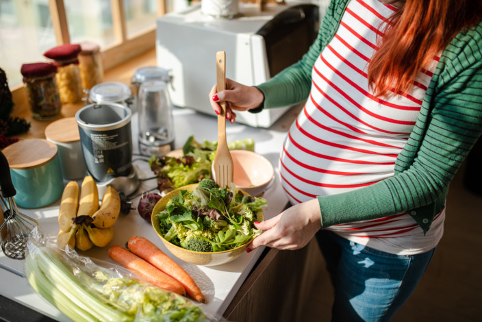 Schwangere bereitet Salat zu auf einer Küchenzeile zu, auf der viel Obst und Gemüse ausliegt