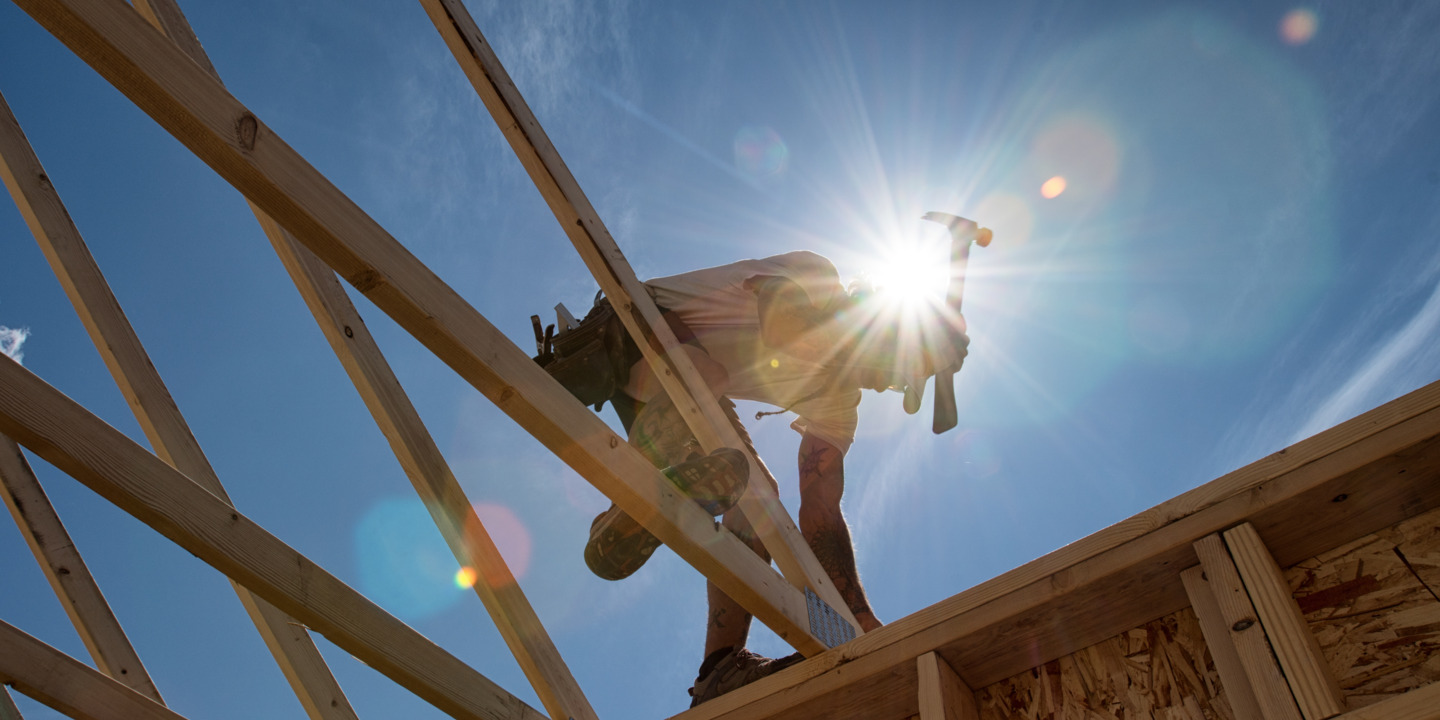 Ein Handwerker arbeitet an dem Dach eines Holzhauses mit einem Hammer in der starken Mittagssonne