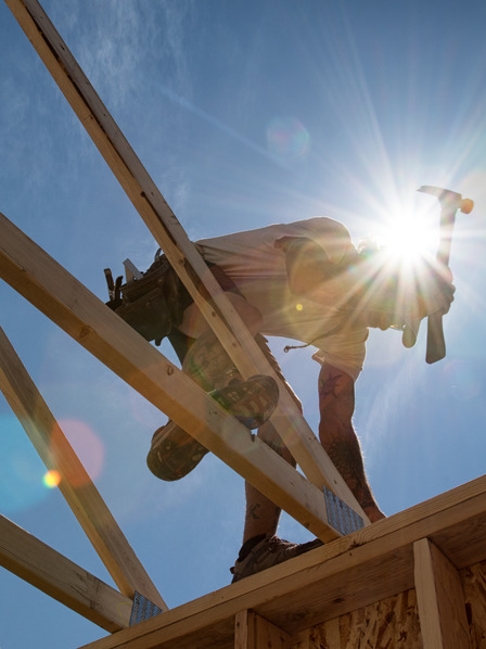 Ein Handwerker arbeitet an dem Dach eines Holzhauses mit einem Hammer in der starken Mittagssonne