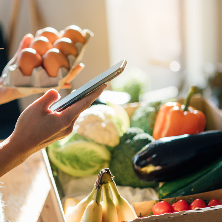 Person mit Smartphone steht vor einer Box mit Obst und Gemüse