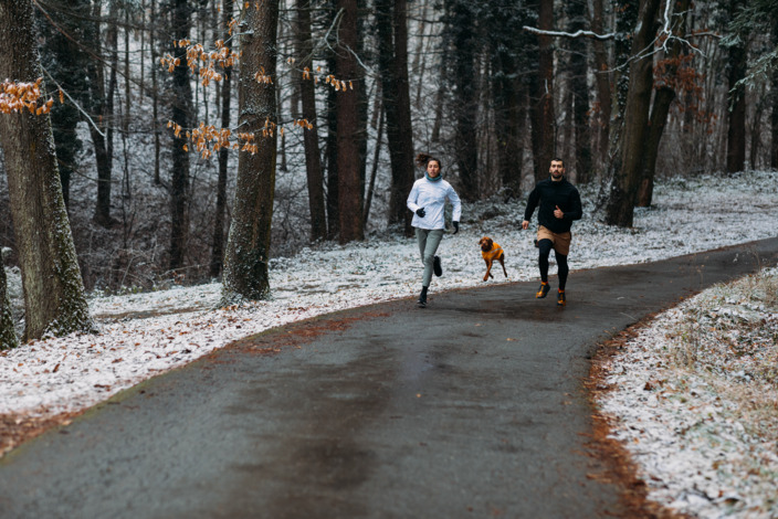 Zwei Personen und Hund joggen im Winter durch den Wald.