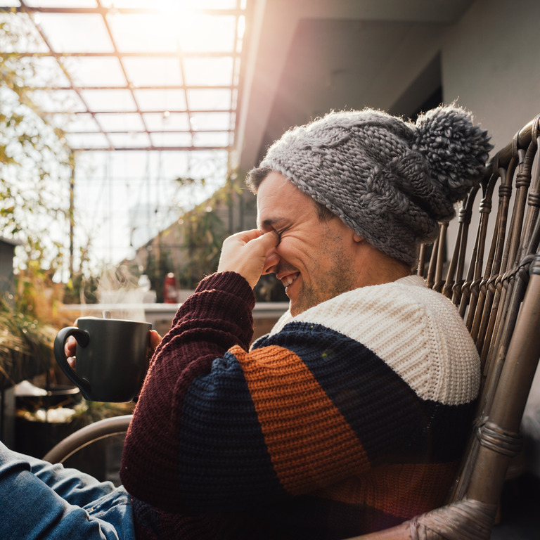 Mann in warmer Kleidung und mit einem Heißgetränk nutzt einen schönen Herbsttag, um auf dem Balkon Sonne gegen das Stimmungstief zu tanken