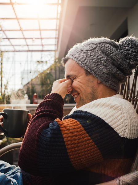 Mann in warmer Kleidung und mit einem Heißgetränk nutzt einen schönen Herbsttag, um auf dem Balkon Sonne gegen das Stimmungstief zu tanken
