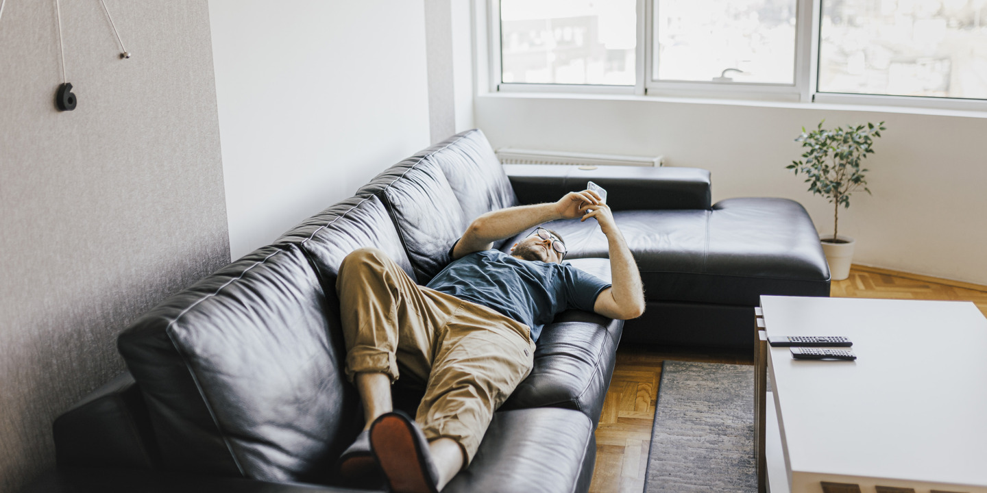 Junger Mann schaut in seine Smartphone auf einer Couch liegend