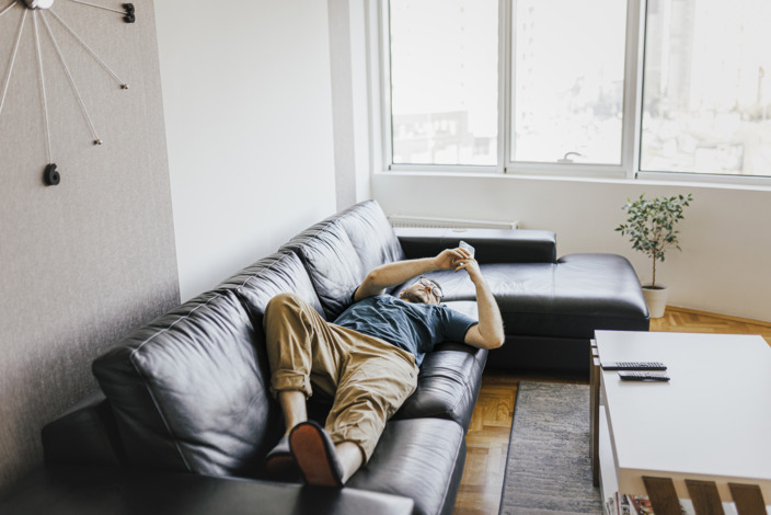 Junger Mann schaut in seine Smartphone auf einer Couch liegend