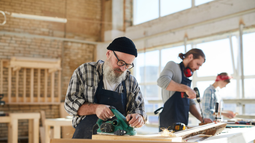 Schreiner im Seniorenalter steht gemeinsam mit jüngeren Kollegen in der Werkstatt und schleift ein Holzstück