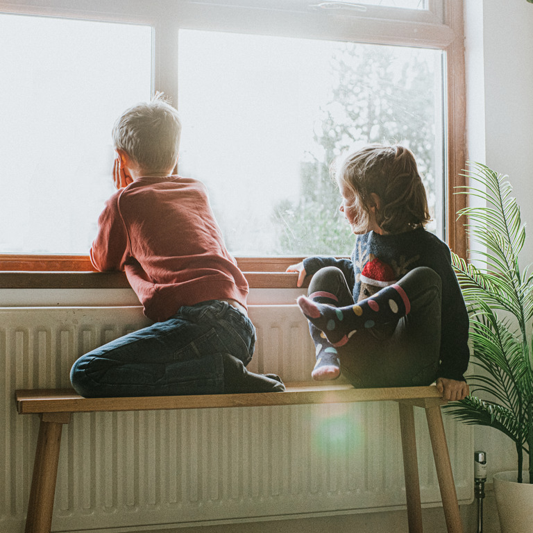 Kinder sitzen in Wohnung an der Heizung und schauen aus dem Fenster