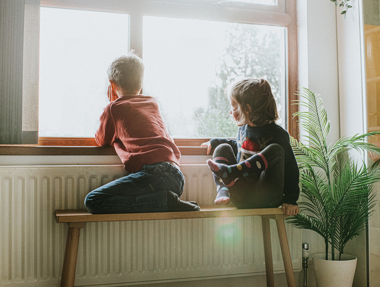 Zwei Kinder sitzen auf einer Bank an der Heizung und schauen aus dem Fenster. 