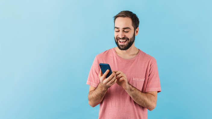 Mann in rosa Shirt vor blauem Hintergrund tippt lächelnd auf dem Smartphone.