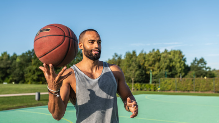 junger Mann steht auf Sportplatz und hält einen Basketball hoch