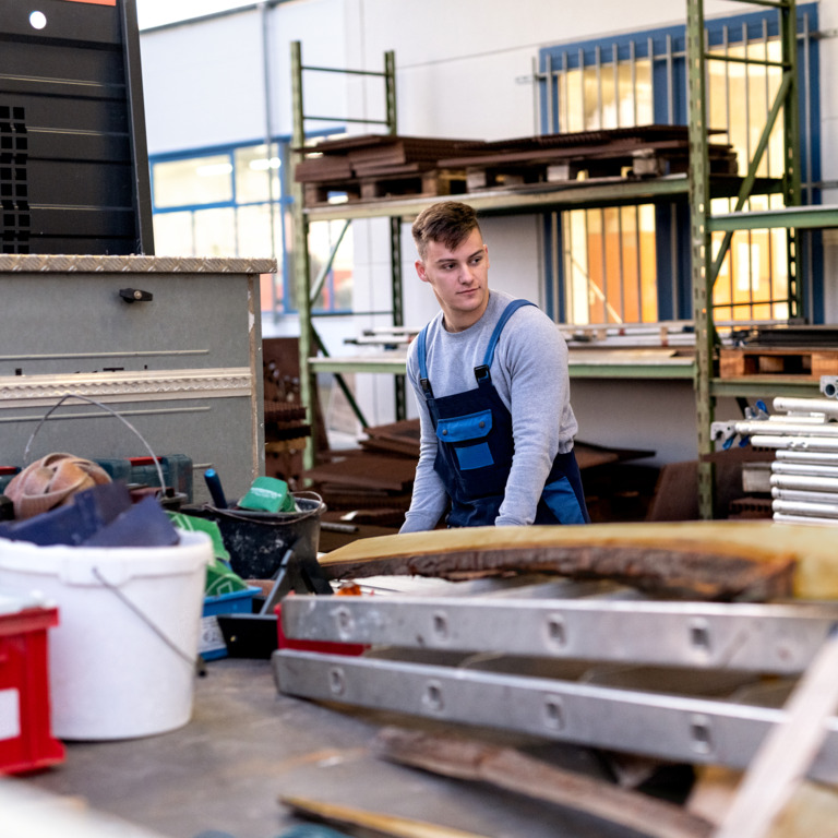 Junger Handwerker in Latzhose steht in Halle und arbeitet.