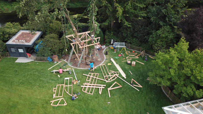 Das Bodengerüst des Baumhauses ist bereits im Baum befestigt, vorgefertige Rahmenteile für Seitenwände und Dachstuhl liegen verteilt auf dem Rasen